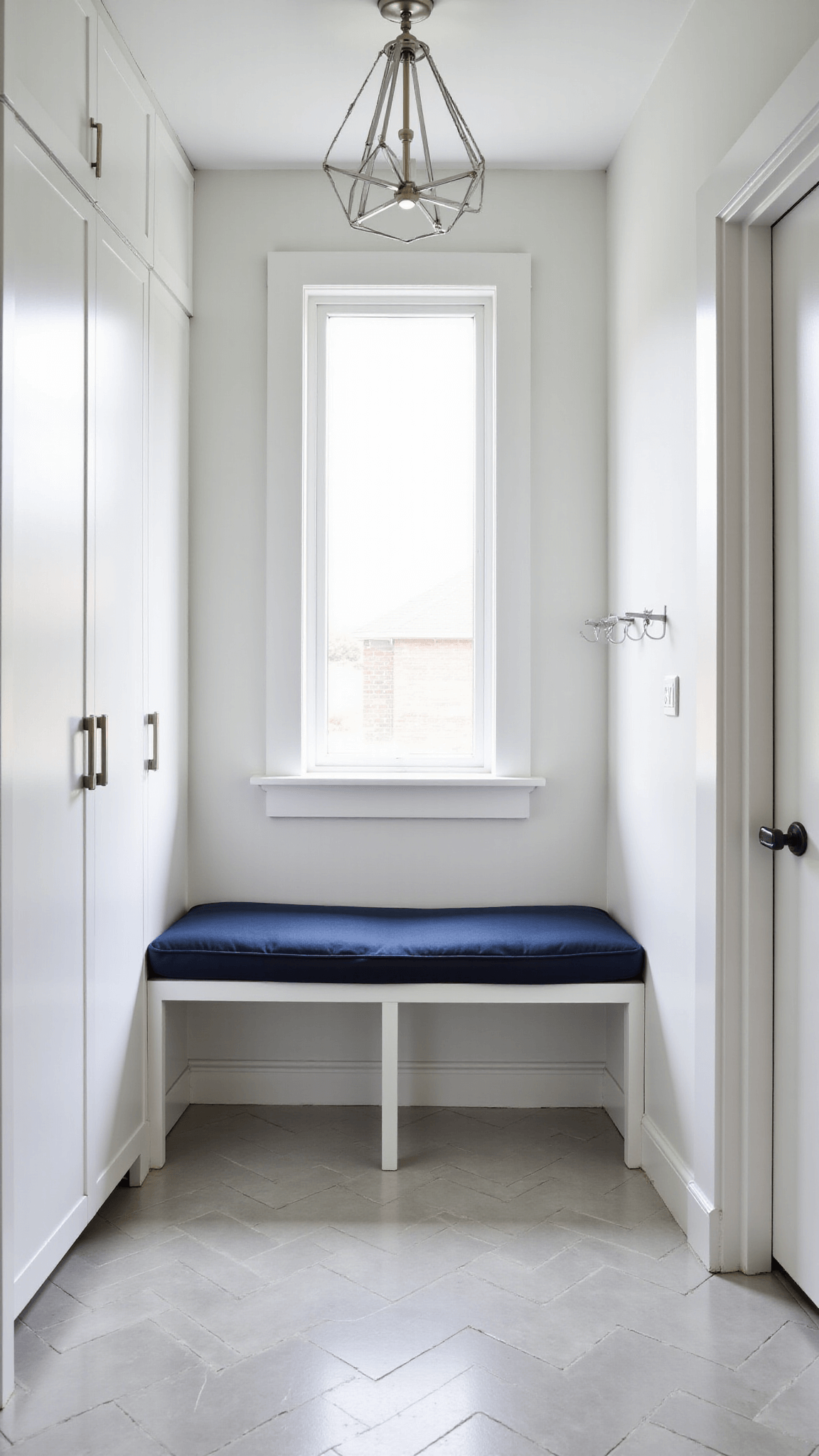 Compact minimalist mud room with white aluminum cabinets, navy cushioned bench, pale gray herringbone tile flooring, modern nickel coat hooks, and geometric pendant light, lit naturally from a transom window.